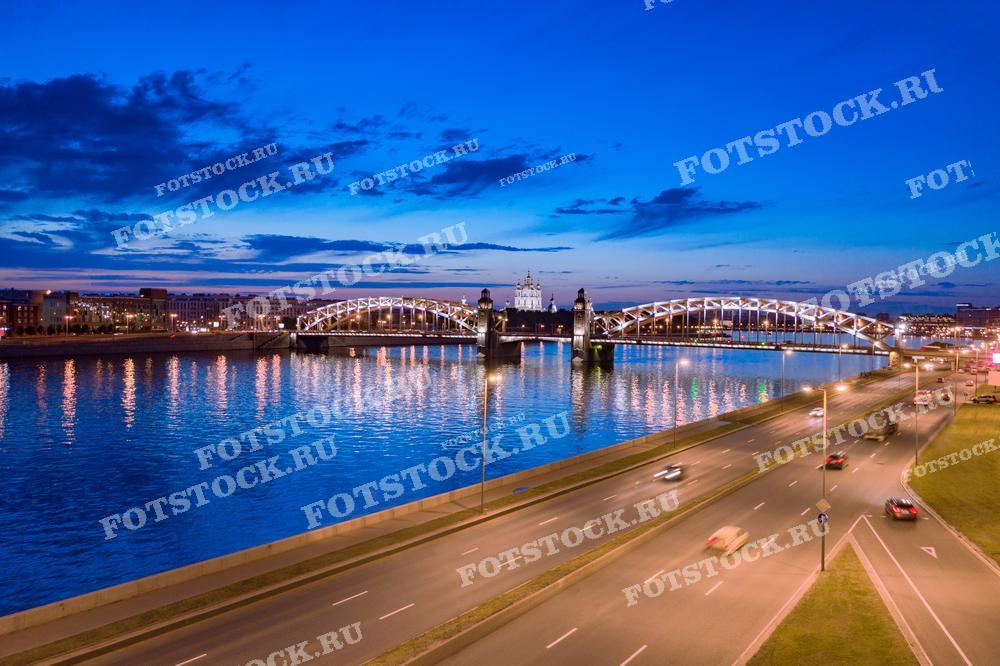 На фотографии запечатлен знаменитый Большеохтинский мост через реку Неву в Санкт-Петербурге в сумеречное время суток. Освещенный мост создает яркую линию через воду, отражение которой видно на водной глади. На заднем плане виднеется Смольный собор с его выразительными синими куполами, освещенными оранжевыми лучами заката. Такой снимок может быть использован для иллюстрации городских гидов, культурно-исторических изданий, туристических материалов, а также в качестве обложки для рекламных брошюр, рассказывающих о красоте и архитектуре Санкт-Петербурга.